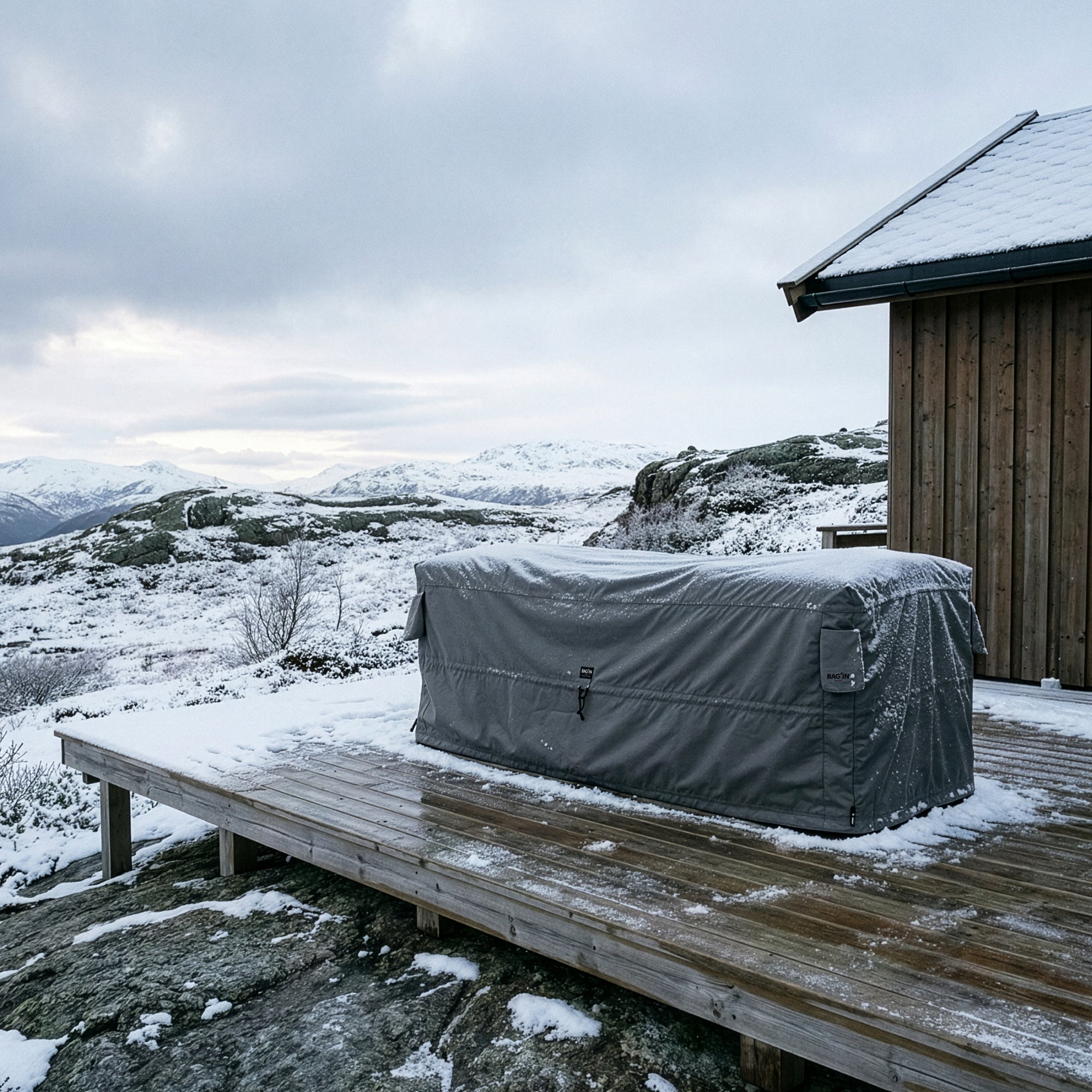 Dark grey BAG’IN Nordic furniture cover protecting outdoor furniture on a wooden deck in snowy mountain conditions.
